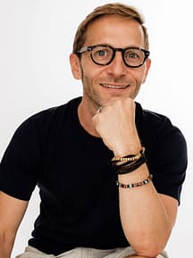 Professional photograph of Francois Souyri (blog author), who is wearing brown glasses and a short sleeves black shirt with the hand on the chin. HE is smiling at the camera and wears bracelets on his left arm.